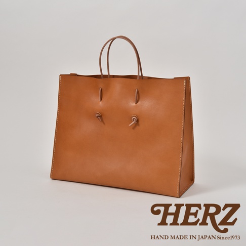 職人手作皮革提袋 T-5，採用棕褐色皮革製成，包身線條簡潔，提把為細長皮革帶，前方有兩個皮革繩結裝飾，側邊及底部有明顯縫線。