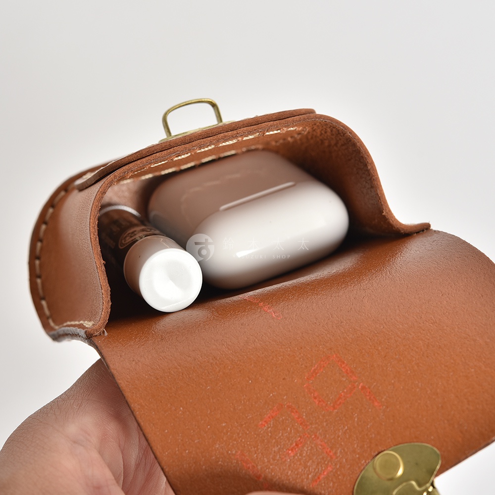 職人手作棕色皮革耳機收納包，內含白色無線耳機充電盒與一小型管狀物品，包蓋以金屬扣固定。