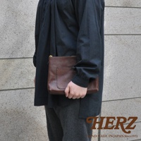 一只手拿著棕色皮革製成的職人手作小物收納包，包上有一個方形外袋，使用金色拉鍊。