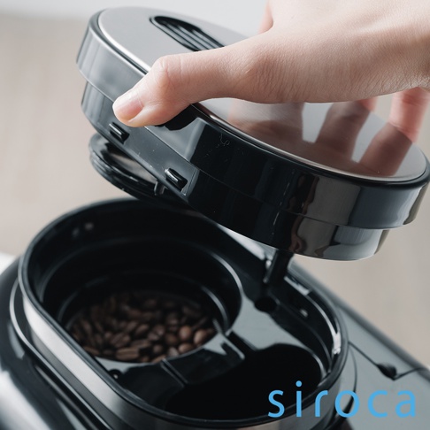 SC-A1210 自動研磨咖啡機，黑色機身，上方為可拆卸的黑色研磨蓋，蓋子頂部為銀色金屬材質。內部裝有淺棕色的咖啡豆。