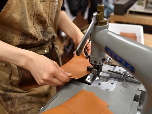 職人雙手拿著咖啡色皮革，在工業縫紉機上進行製作。縫紉機為灰色，帶有「KOPRO」字樣。
