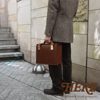 棕色皮革機師箱，配有金色鎖扣和手柄，由一位身穿格紋外套的男士手持，背景為戶外階梯與建築。