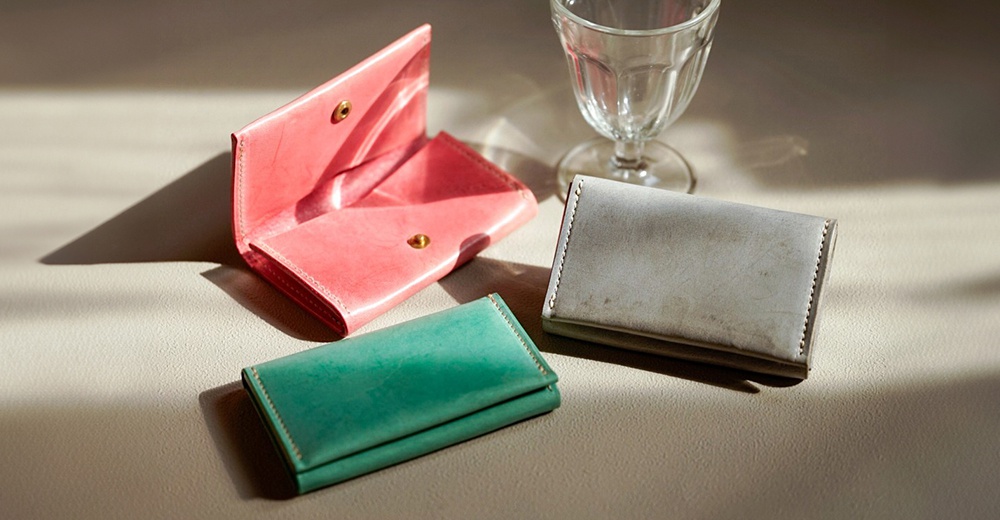 北海道KASANE三款皮革卡夾，分別為粉紅色、綠色及灰色，擺放在淺色紋理背景上，並有自然光線投下的陰影，旁邊還有一只透明玻璃杯。