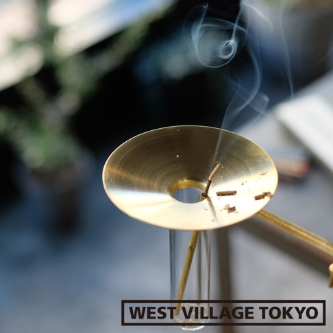 黃銅製的圓盤狀線香架，中央有孔，用於固定線香，線香燃燒產生藍色煙霧，線香架下方為透明玻璃管，整體呈金屬光澤。