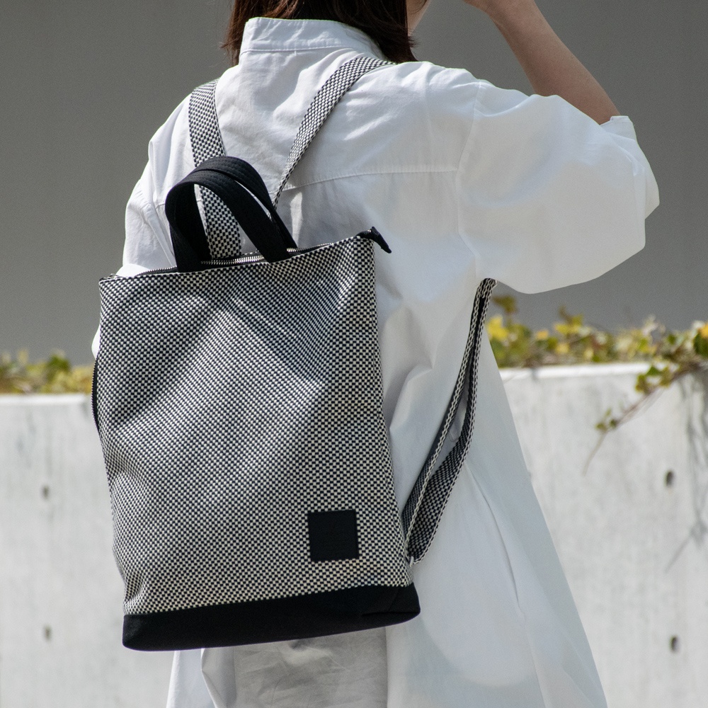 一款三河木綿刺子織手提後背包，以黑白小格紋圖案為主體，配有黑色提把與底部，另有黑色方形布標裝飾。