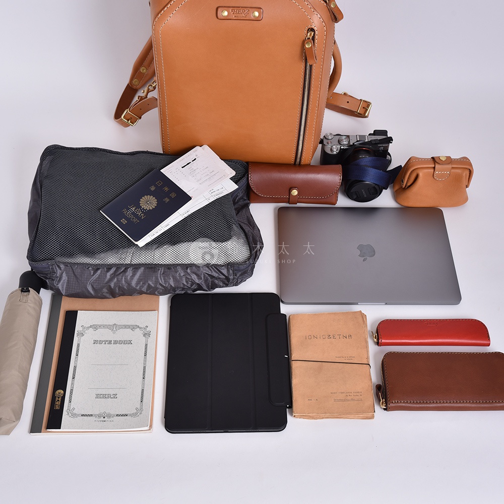 一張展示職人手作皮革兩用商務後背包 R-33 的圖片，背包為棕褐色皮革。周圍擺放著電腦、平板、相機、筆記本、護照、文件夾、雨傘及各式皮革小物。