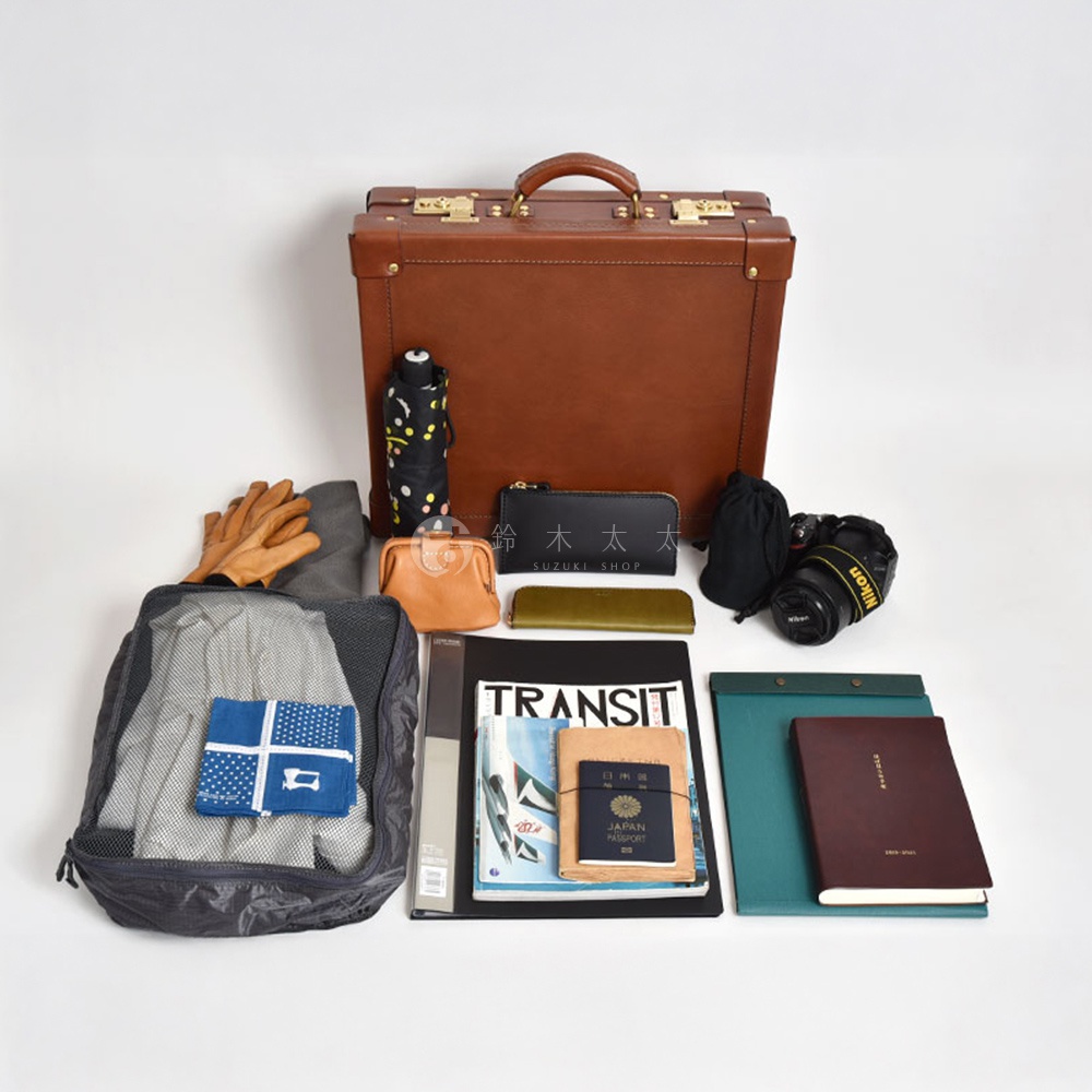 棕色皮革手提行李箱，搭配金色釦飾。箱旁有黑色印花雨傘、皮革手套、橘色零錢包、黑色長夾、綠色筆、黑色相機、灰色收納袋，內含衣物及藍色印花手帕，以及一本《TRANSIT》雜誌、一本日本護照和一本深紅色筆記本。