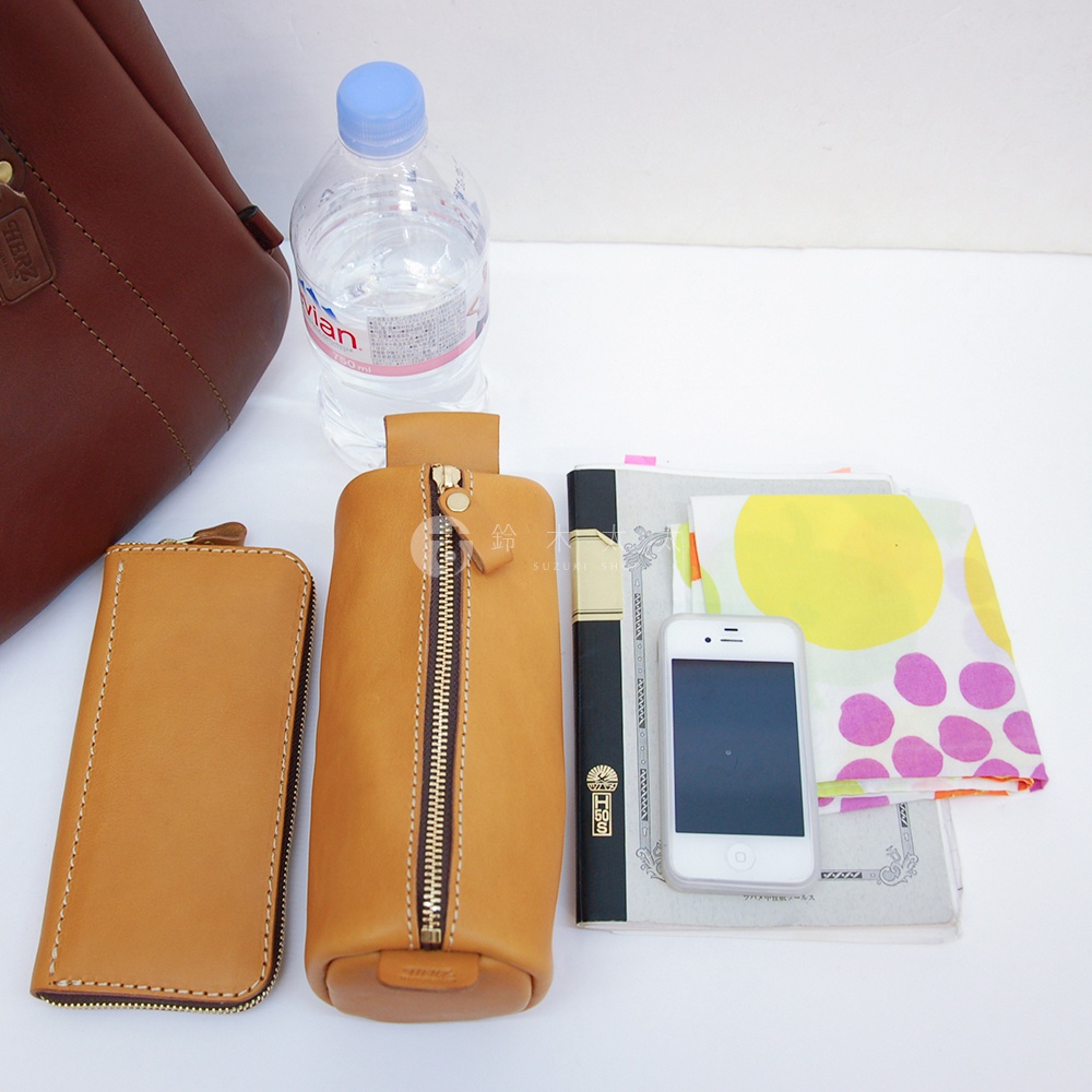 職人手作皮革仕女托特包T-10，展示了棕色皮革長夾、淺棕色皮革圓筒筆袋、一瓶礦泉水、一本黑色筆記本、一本筆記本和一個白色iPhone。