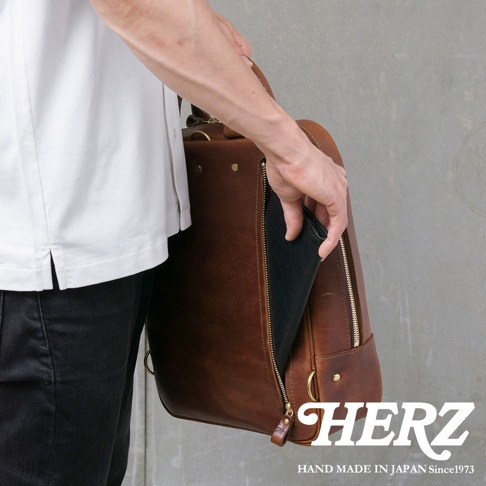 一人伸手將一個黑色皮夾放入一個棕色皮革背包的拉鍊側袋，背包上有HERZ字樣和HAND MADE IN JAPAN Since1973字樣