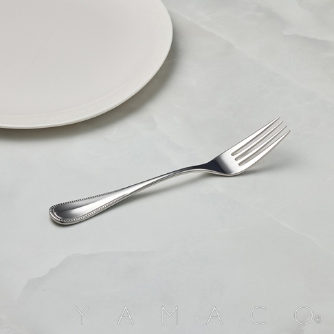 一個不鏽鋼叉，有著珠狀裝飾的握柄，放置在一個白色盤子旁邊。