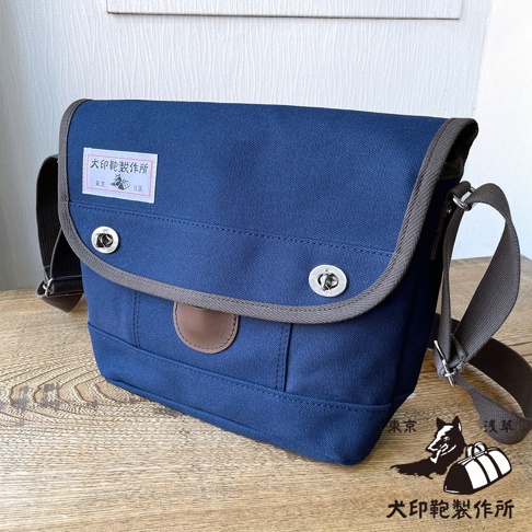 海軍藍純綿帆布雙扣郵差包，配有灰色滾邊和棕色皮革裝飾，正面中央有兩個銀色旋轉鎖扣，左側有一個印有品牌標誌的白色方形標籤。