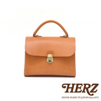 日本HERZ手工皮革包，焦糖色，翻蓋磁扣設計，頂部有提把，下方有HERZ品牌標誌與「HAND MADE IN JAPAN Since1973」字樣