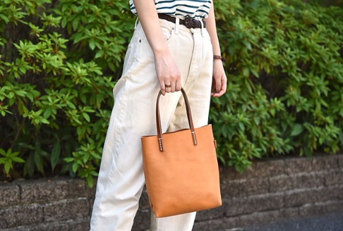 一名女子手持一個淺棕色皮革直式軟皮托特包，包包上有深棕色皮革提把，並以白色縫線加固。