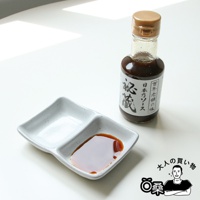 玻璃瓶裝著深棕色醬汁，瓶身貼有印有日文的白色標籤。一旁有一個雙格的淺灰色陶瓷醬料碟，其中一格盛有少量醬汁。