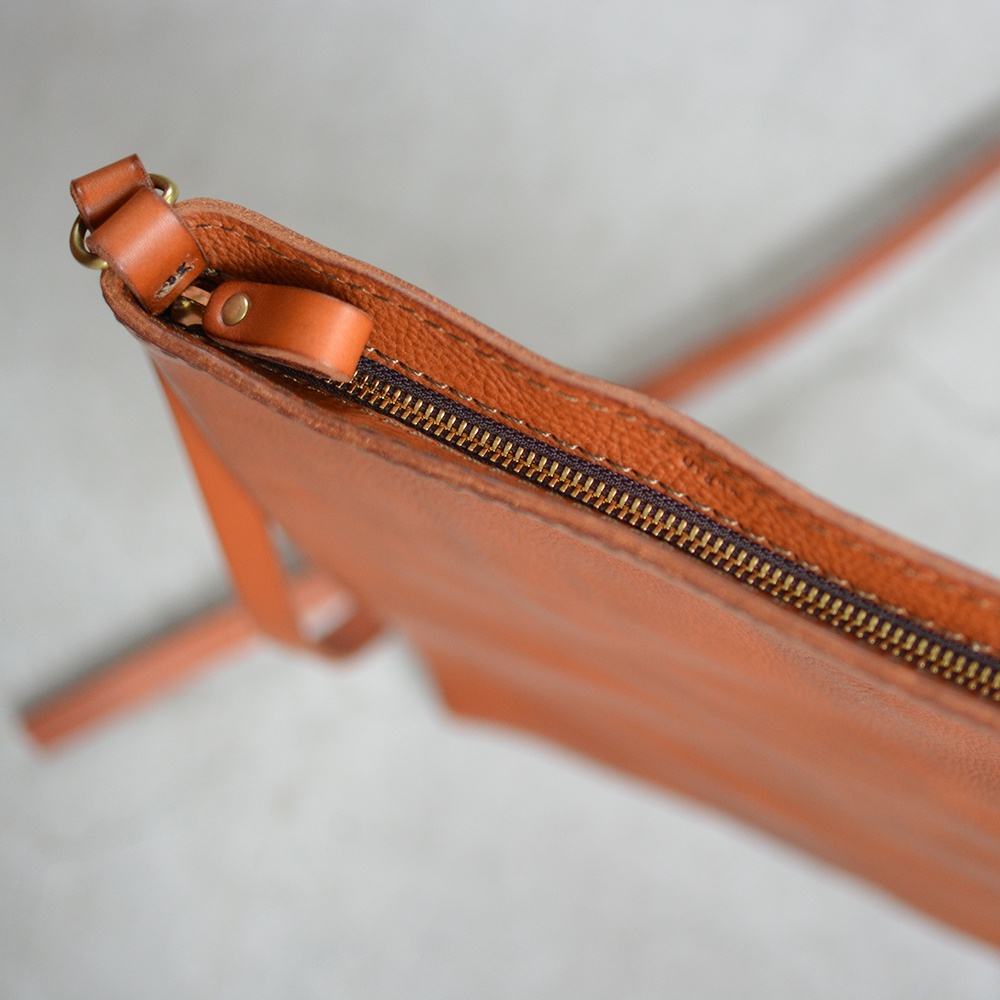 橘棕色皮革材質的肩背包，特寫拉鍊細節與皮革縫線，附有簡約的皮革背帶。