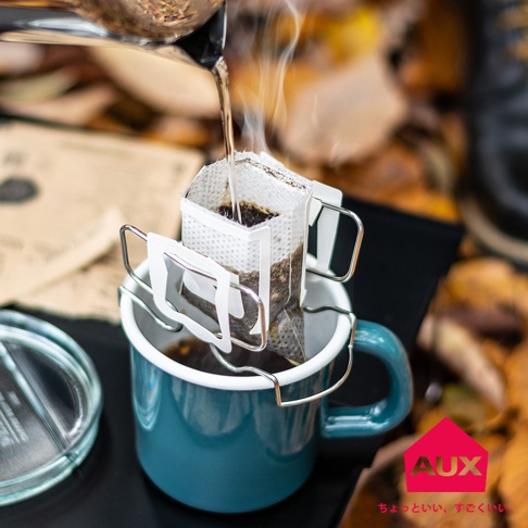 銀色金屬可調式耳掛咖啡架，夾著一個白色濾紙包，正在滴漏咖啡液至藍色馬克杯中，杯中已有部分咖啡。