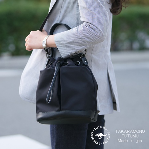 一個斜挎著黑色抽繩水桶包的人，包上有TAKARA MONO TUTUMU字樣，包底部有皮革裝飾。