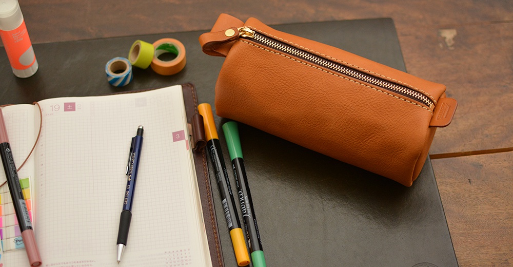 職人手作皮革多用途收納筆袋，採用棕褐色皮革製成，配有金色拉鍊和提把。筆袋旁有筆記本、原子筆、馬克筆和彩色膠帶。