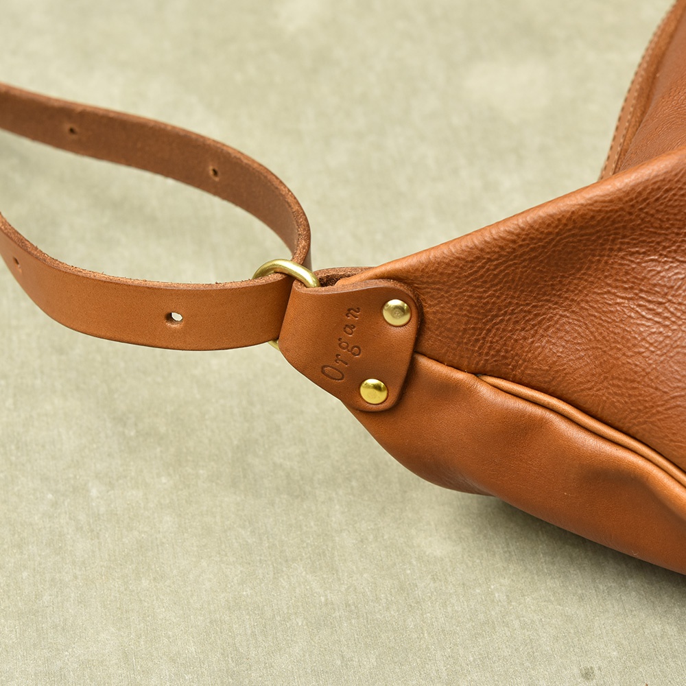 職人手作皮製單肩半月包，採用棕色皮革，帶有品牌壓紋和黃銅色五金配件，搭配可調節的皮質肩帶。