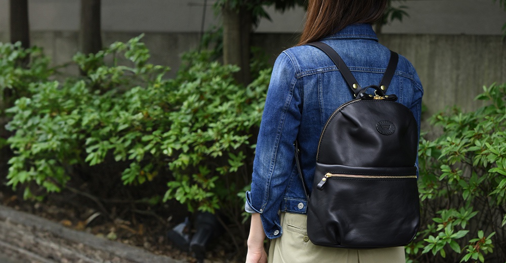 黑色皮革大開口圓角後背包，配有金色拉鍊和品牌壓紋。