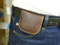 職人手作皮革馬蹄造型零錢包，深棕色皮革，縫線細緻，搭配牛仔褲與皮帶，展現復古風格。