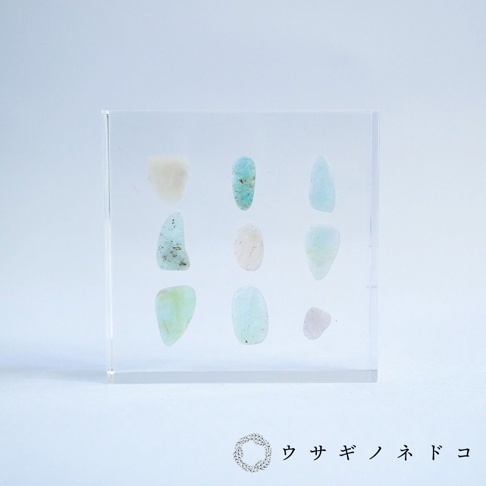方形透明壓克力塊中，整齊排列著九顆大小不一、形狀各異的細石。這些細石呈現多種柔和色彩，包含淺白、淡藍、淡綠及粉紅，材質看起來像玉石或半寶石。