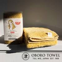 一條黃色紋理柔軟的專用髮巾，以透明包裝盒展示，旁邊疊放著兩條同樣材質的黃色毛巾。