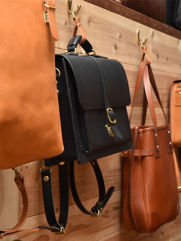 職人手作皮革吊掛扣帶 (KZ-3)。黑色皮革背包，搭配金屬扣環，懸掛於木質牆面上，旁邊還有棕色皮革包。