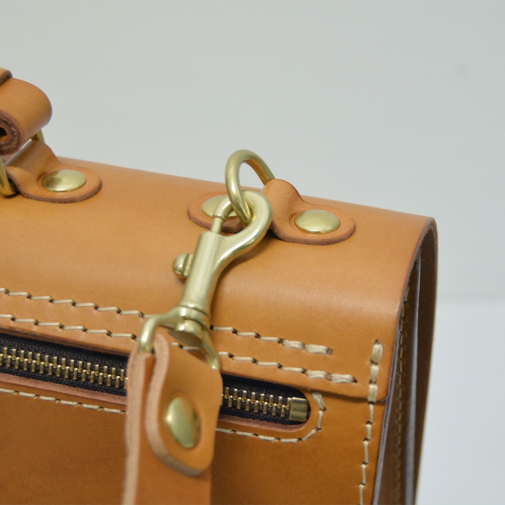 職人手作皮革旅行商務兩用波士頓包V-3，此圖特寫其淺棕色皮革材質，搭配古銅色金屬五金配件，包括側邊的拉鍊、提把固定鈕扣及可拆卸肩帶的鉤環，展現精緻的縫線工藝。