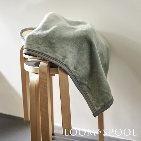 淺綠色短絨舒柔單人毯，表面呈現細緻絨毛感，邊緣有深灰色滾邊，毯子披掛在木製圓凳上。