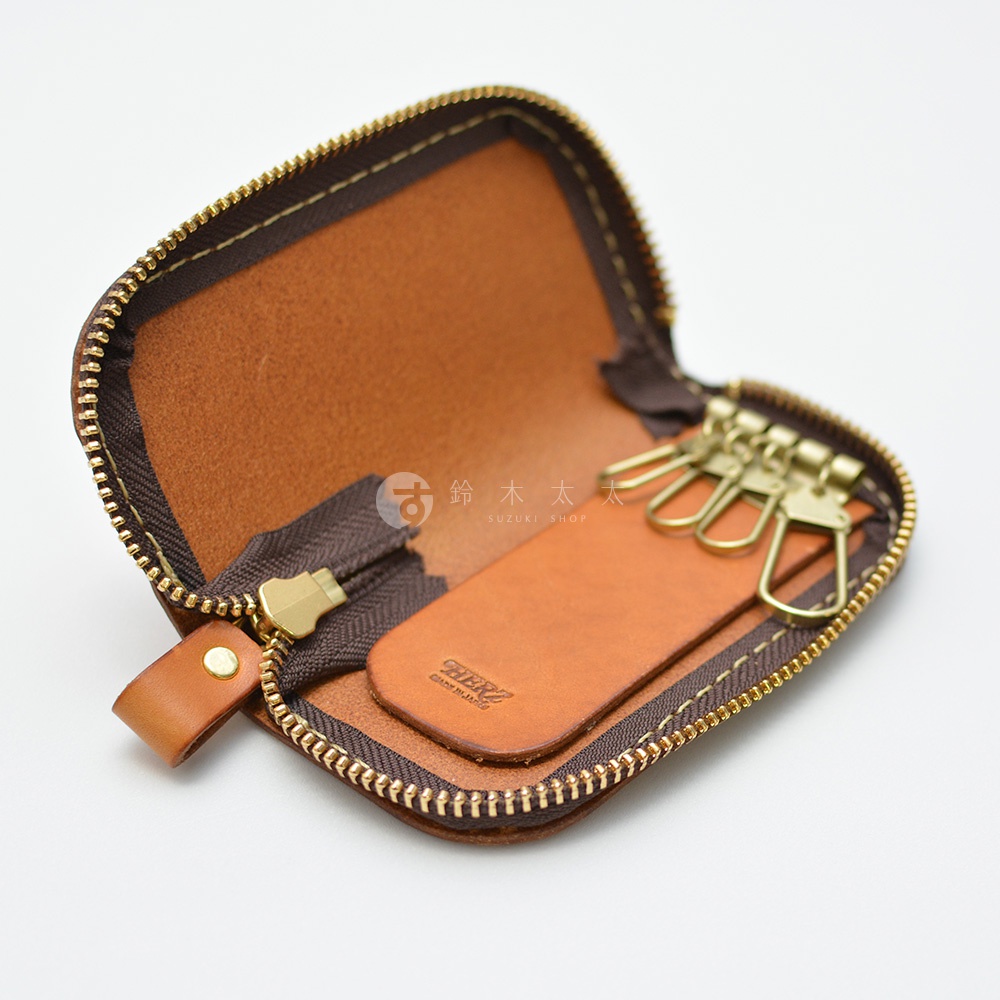 職人手作皮革橢圓形拉鍊鑰匙包，內部設有四個金屬掛鉤，皮革上印有品牌標誌。