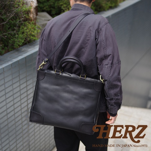 男士身背黑色皮革托特包，包包有雙提把和可拆卸肩帶，採用厚實皮革製作，設計簡約。