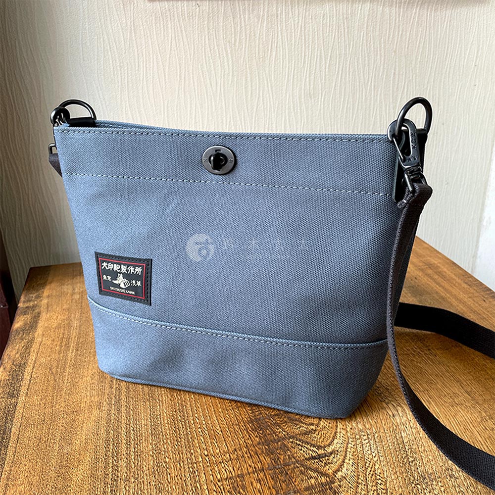 犬印鞄製作所的灰色帆布迷你側背包，上面有品牌標籤，包包上有金屬扣環和黑色背帶，放置在木質桌面上。