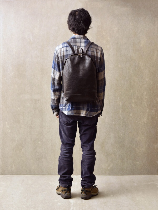 一名男性身穿藍色格紋襯衫、深色長褲，背著一個深棕色皮革後背包。後背包呈現簡約的褶皺風，頂部有兩個拉鍊。