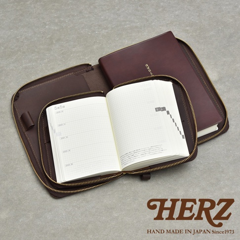深棕色皮革記事本收納包，拉鍊部分打開，露出內頁有格子和文字的記事本，右側為封閉的記事本，下方有HERZ品牌標誌。
