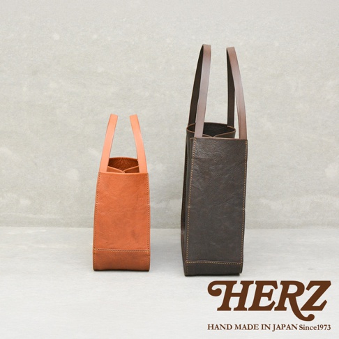 兩個不同尺寸與顏色的日本HERZ手工皮革托特包並排擺放，左側為淺棕色的小型包款，右側為深棕色的大型包款，兩者皆具備簡約的方形立體結構與長提把設計，底部右下方印有HERZ品牌名稱與相關標語。