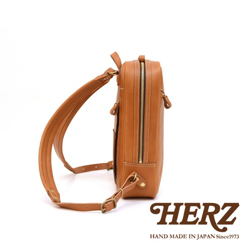 職人手作皮革長方型後背包，展示為焦糖棕色，採用高品質皮革製作，側邊可見拉鍊與外口袋設計，搭配精緻縫線與金屬扣環，風格簡約實用。