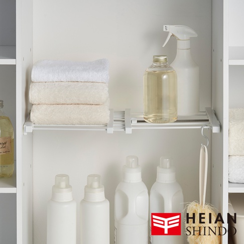 一個寬版伸縮縫隙置物架，上面擺放著疊起的毛巾、一瓶透明液體、一個白色噴瓶，下方則是幾個白色瓶狀清潔劑和一個洗澡刷。