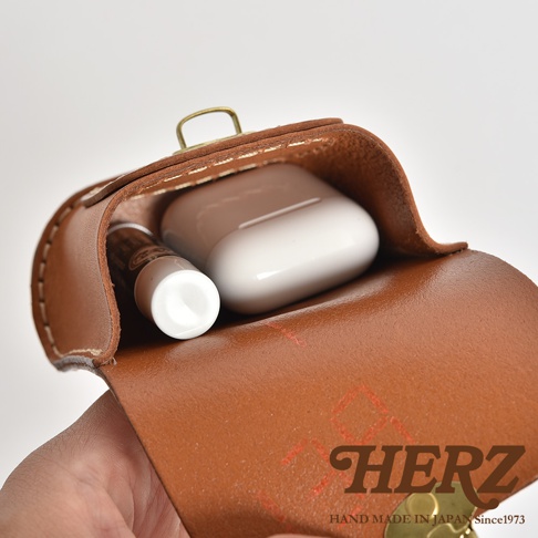 棕色皮革耳機收納包，內含白色無線耳機及一小瓶狀物，以黃銅釦固定，邊緣縫線清晰，材質為牛皮。
