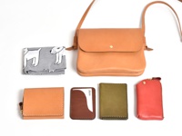 職人手作皮革橫式隨行袋 (CP-9)，搭配卡其色皮革短夾、棕色皮革卡夾、墨綠色皮革名片夾及紅色皮革鑰匙包。另有一塊灰色印有白色狗狗圖案的布料。