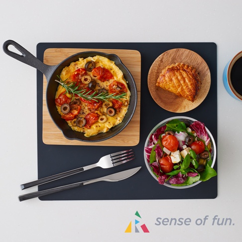 黑色耐高溫微波爐防污保護墊上，擺放著一個鑄鐵鍋，鍋中盛有番茄、橄欖和迷迭香裝飾的烘蛋；旁邊有一個木盤，上面放著一個酥皮派；下方有一個透明碗，裝有番茄、奶酪和綠葉蔬菜組成的沙拉；另外還有銀色金屬叉和刀，手柄為黑色。