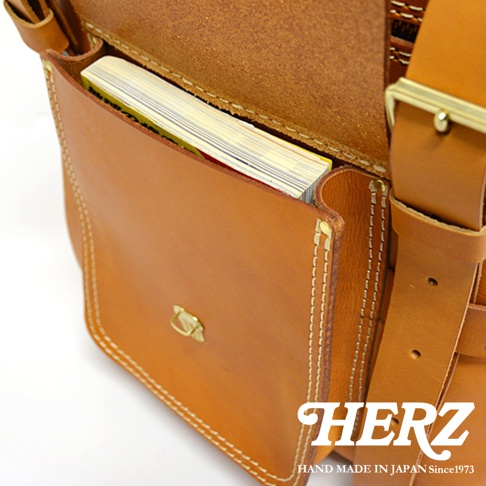 職人手作皮革大型三用旅行後背包 A-21，展示了棕褐色皮革的正面翻蓋與側邊。包包內側可見一本黃色封面的書本，翻蓋處有金屬釦環，縫線清晰。