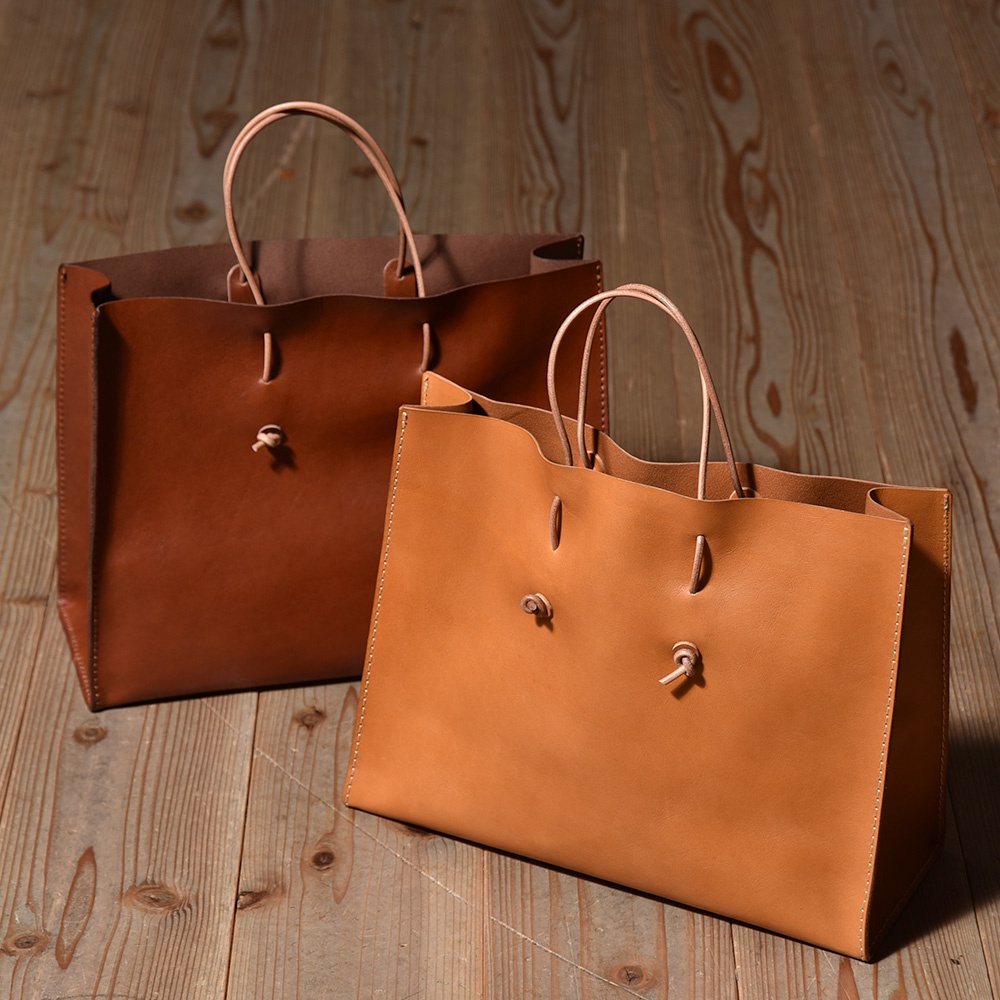 兩款職人手作皮革提袋，一款為深棕色，另一款為淺棕色。兩者皆為大地色系，材質為皮革，設計簡約，提把為圓形，袋口處有皮革繫繩設計。