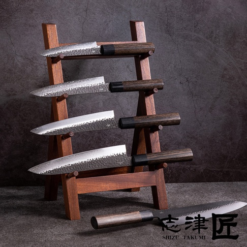 木質刀架上擺放著四把銀色金屬刀刃、深棕色木質手柄的主廚刀，刀刃帶有錘紋和 Damascus 紋理。其中一把刀平放在前景，刀柄朝左。