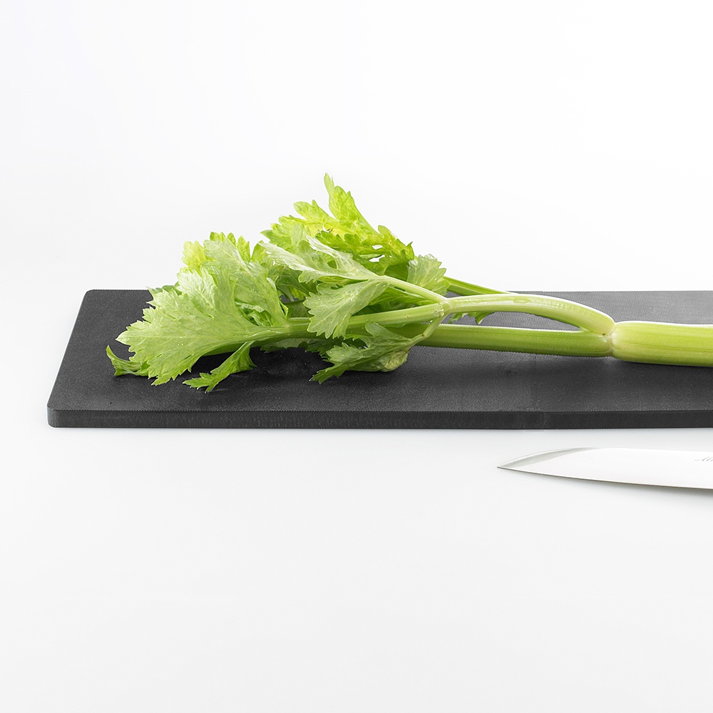 黑色砧板上放著一把刀，旁邊是新鮮的芹菜，葉子呈淺綠色，莖部較粗。