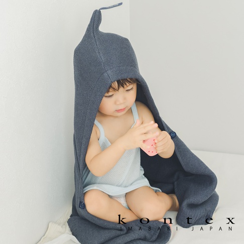 一位穿著淺藍色內衣的嬰兒，被一件深灰色針織風純棉連帽浴巾包裹著。浴巾頂部有一個尖角造型，並附有品牌標籤。