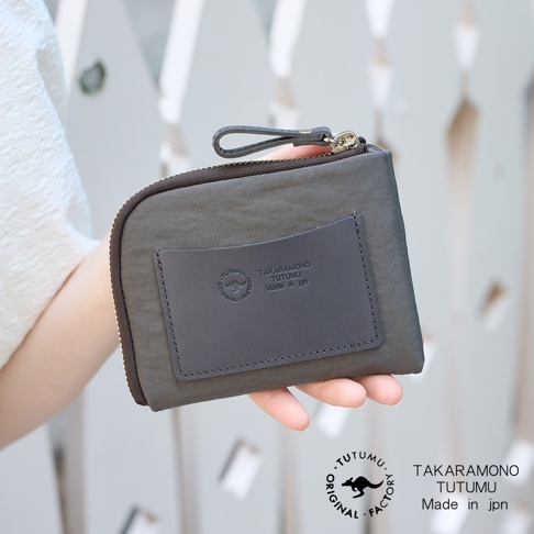 TUTUMU 品牌防潑水迷你錢包，以手持方式呈現，錢包主體為灰色，帶有深灰色的皮革標籤，上面印有品牌標誌和產地資訊「TAKARA MONO TUTUMU Made in jpn」，右側有拉鍊開合，左側有織物材質。