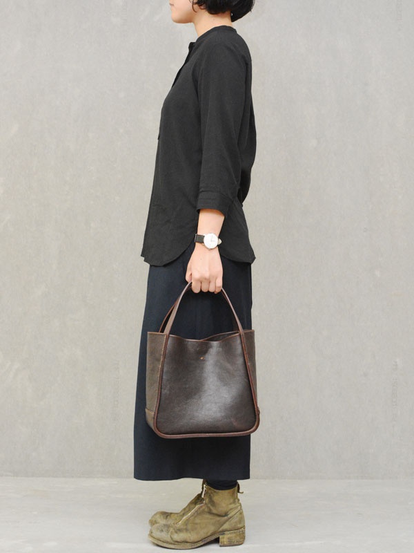 一位女士手持深棕色皮革方形托特包，包包有兩個提把，包身結構簡潔，皮革表面呈現自然的紋理，整體風格樸實。