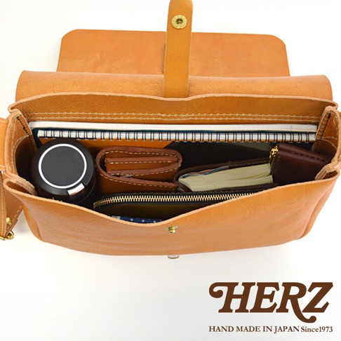 日本HERZ手工製作的棕色皮革軟皮橫式斜背包，內部裝有筆記本、錢包和圓形黑色小物，側邊有品牌標誌。