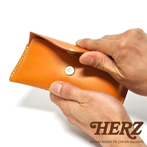 職人手作皮革眼鏡收納盒，橘褐色皮革材質，以雙手操作開合，品牌HERZ標誌與MADE IN JAPAN字樣清晰可見。